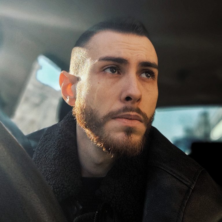 A man with a beard sits in a car, looking thoughtfully out the window with sunlight on his face.